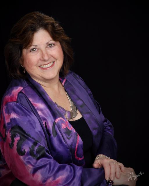 In a studio portrait setting, Deb smiles broadly with her hands crossed in front of her. She&#039;s wearing a purple silk jacket. 