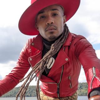 Image of a person wearing a red leather jacket and red hat cocked sideways. They have long dreads and are reaching toward the camera. Jé Exodus Hooper (they/them) a non-binary preacher who is clergy at First Unitarian Society of Minneapolis 
