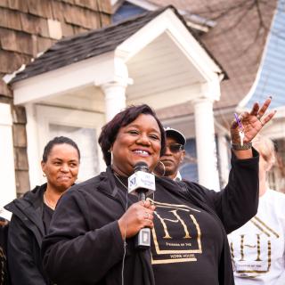 An African American woman in an all-black outfit holds a set of house keys. She is standing in front of a multiracial group of people in the front yard of a house.