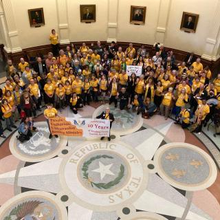 A multiethnic, multi-gender group of people in yellow shirts/tops in a foyer of a state building. The floor reads "Republic of Texas".