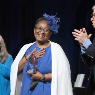 A smiling person in a blue dress, white shawl and a matching blue fascinator hat receives a modern glass award for distinguished service to Unitarian Universalism as people clap.