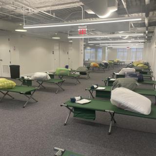 Rows of green cots in an open office space with white walls and gray carpet. The cots have bundles on them.