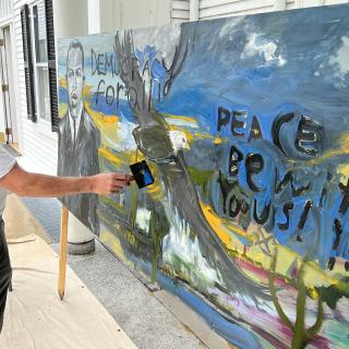 A person creating a large painting during a democracy festival. The painting features Civil Rights icon John Lewis, a bald eagle and the words "peace be with you, us."