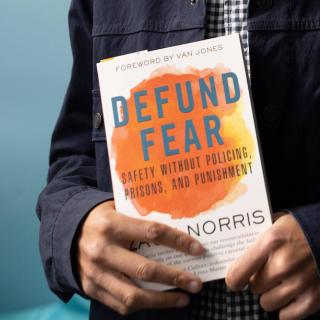  book titled "Defund Fear" by Zach Norris is held in the hands of someone whose hands are light brown; only their torso is showing.