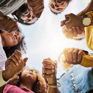 Stock photo of a group of black folx holding hands praying in a circle, photo taken from below. 
