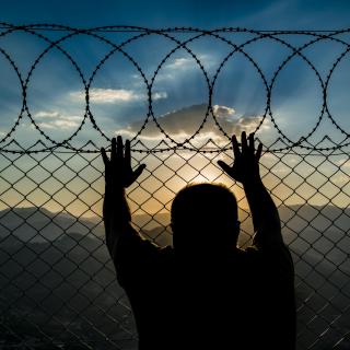 Stock photo of silhouette of person leaning against a chainlink fence topped with razor wire, looking out.