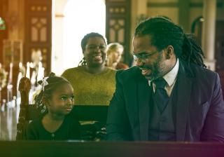 A Black father and young daughter looking happy at church with a smiling Black woman sitting in the row behind them. 