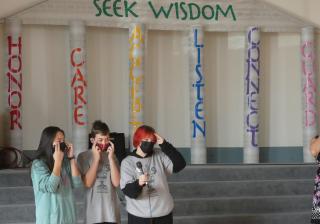 Three young people in face masks due to COVID are standing in front of a greek temple facade. The top of the facade reads "seek wisdom" and the columns read "honor, care, accept, listen, connect, guard."