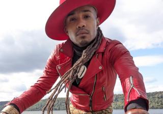 Image of a person wearing a red leather jacket and red hat cocked sideways. They have long dreads and are reaching toward the camera. Jé Exodus Hooper (they/them) a non-binary preacher who is clergy at First Unitarian Society of Minneapolis 