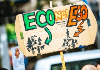 Protest sign reads "Eco not Ego," and depicts man living in harmony with nature, versus man ruling over nature from the top of a pyramid.