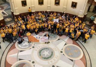 A multiethnic, multi-gender group of people in yellow shirts/tops in a foyer of a state building. The floor reads "Republic of Texas".