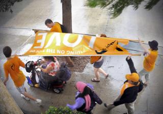 people hold a Side With Love banner over a person in a scooter while moving down a sidewalk in the rain