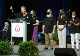 A group of people in matching black shirts stand on stage. In front of them is a smiling person at a lectern, also facing the audience.