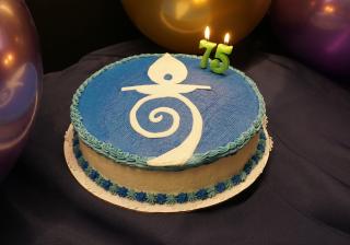 Photograph of a birthday style cake with the logo of the Church of the Larger Fellowship on the top, and 2 lit candles spelling "75". 