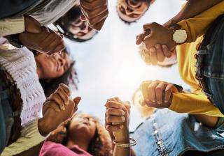 Stock photo of a group of black folx holding hands praying in a circle, photo taken from below. 