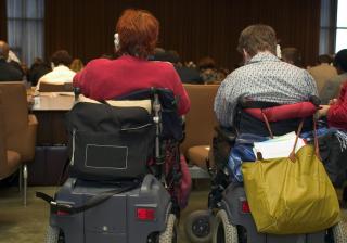 photograph of 2 people in accessibility scooters, in the audience at a conference