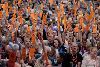 a large indoor gathering holds up orange paper voting cards