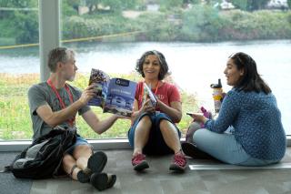 Three people sit on the floor in front of a large window looking at a GA program book