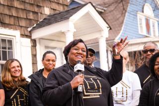 An African American woman in an all-black outfit holds a set of house keys. She is standing in front of a multiracial group of people in the front yard of a house.