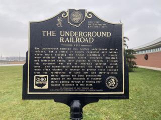 A historic heritage sign about the Underground Railroad. Behind it is a green field, with a brick building to the right.
