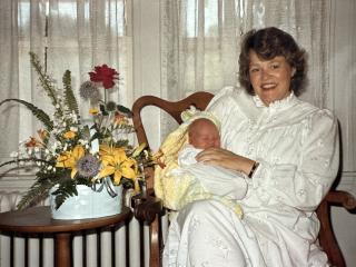 A seated white woman in all-white garb smiling and holding a newborn. To their right is a table on which there are flowers.