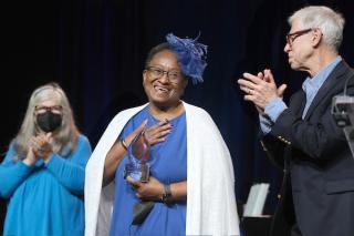 A smiling person in a blue dress, white shawl and a matching blue fascinator hat receives a modern glass award for distinguished service to Unitarian Universalism as people clap.