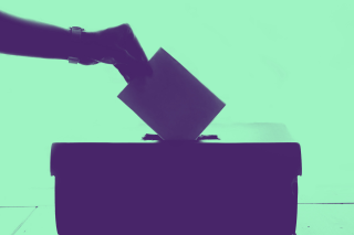 A hand places a paper ballot into a voting box.