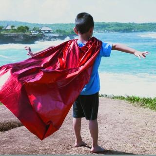 Stock phonto of a boy standing on a rocky outcrop near the ocean, wearing a cape with his arms outstretched.