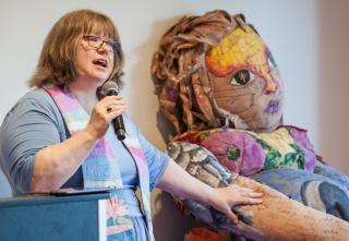 The Rev. Kimberley Debus preaches at a UU Women's Federation event on finding wholeness in a culture of misogynist brokenness, beside a ritual object called Big Woman (created in 1999 by Elizabeth Schell) at GA 2019 