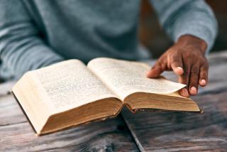 Stock photo of brown hands turning pages of an old book that looks like the bible. 