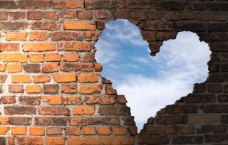 Photo illustration of old brickwork with a hole in the form of a heart, showing blue sky.