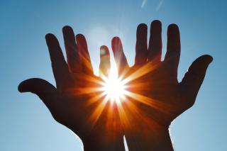 Stock photo of the sun peeking through two silhouetted hands. Blue sky background.