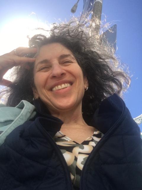 This is a shoulders-up photo of Susan Dana Lawrence, a white woman with salt and pepper wavy hair, smiling into the sun. She is wearing a print shirt, a black sweater, and light blue, collared jacket.