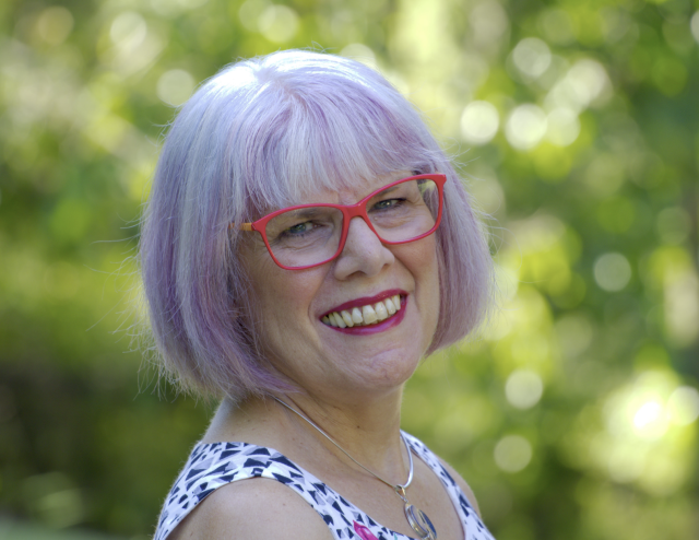 A smiling person with lilac and blue bob and hot pink glasses stands before a green outdoor background.