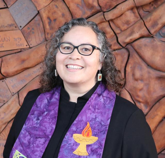 Rev. Tania, wearing a black clergy robe and purple stole, smiles in front of a textured brick wall.