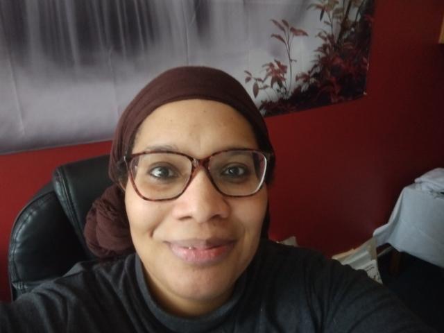 Erica, with light brown skin, wears glasses and a brown head wrap. She smiles in front of a red wall.