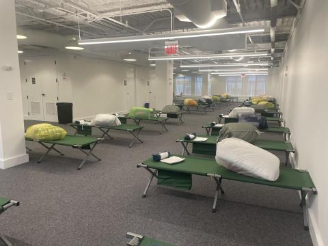 Rows of green cots in an open office space with white walls and gray carpet. The cots have bundles on them.