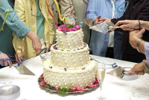 A three-tiered wedding cake with pink flowers on a table. People around it prepare to cut it together.